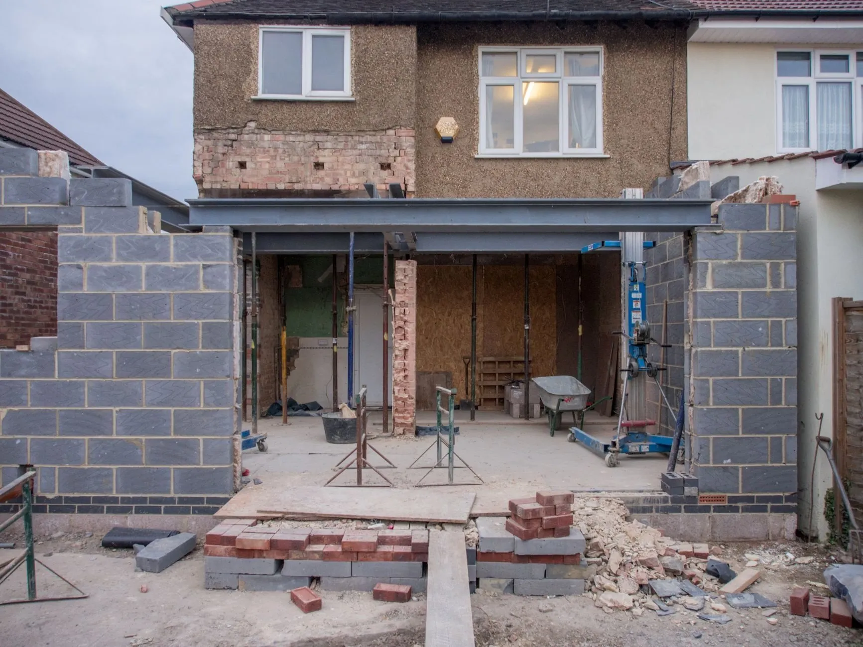 The back of a semi-detached house opened up for a single-storey extension, with steel beams, props and stacked bricks