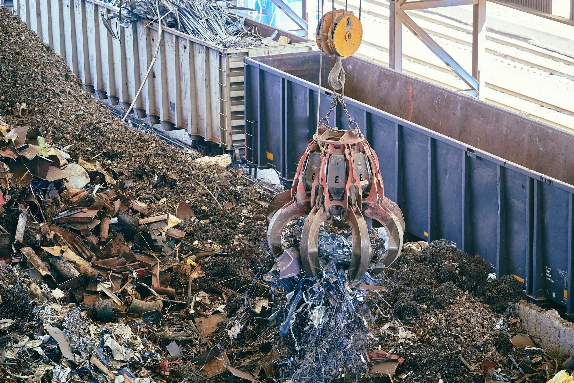 A crane grab lifting scrap metal from a heap beside open steel wagons in a sorting hall