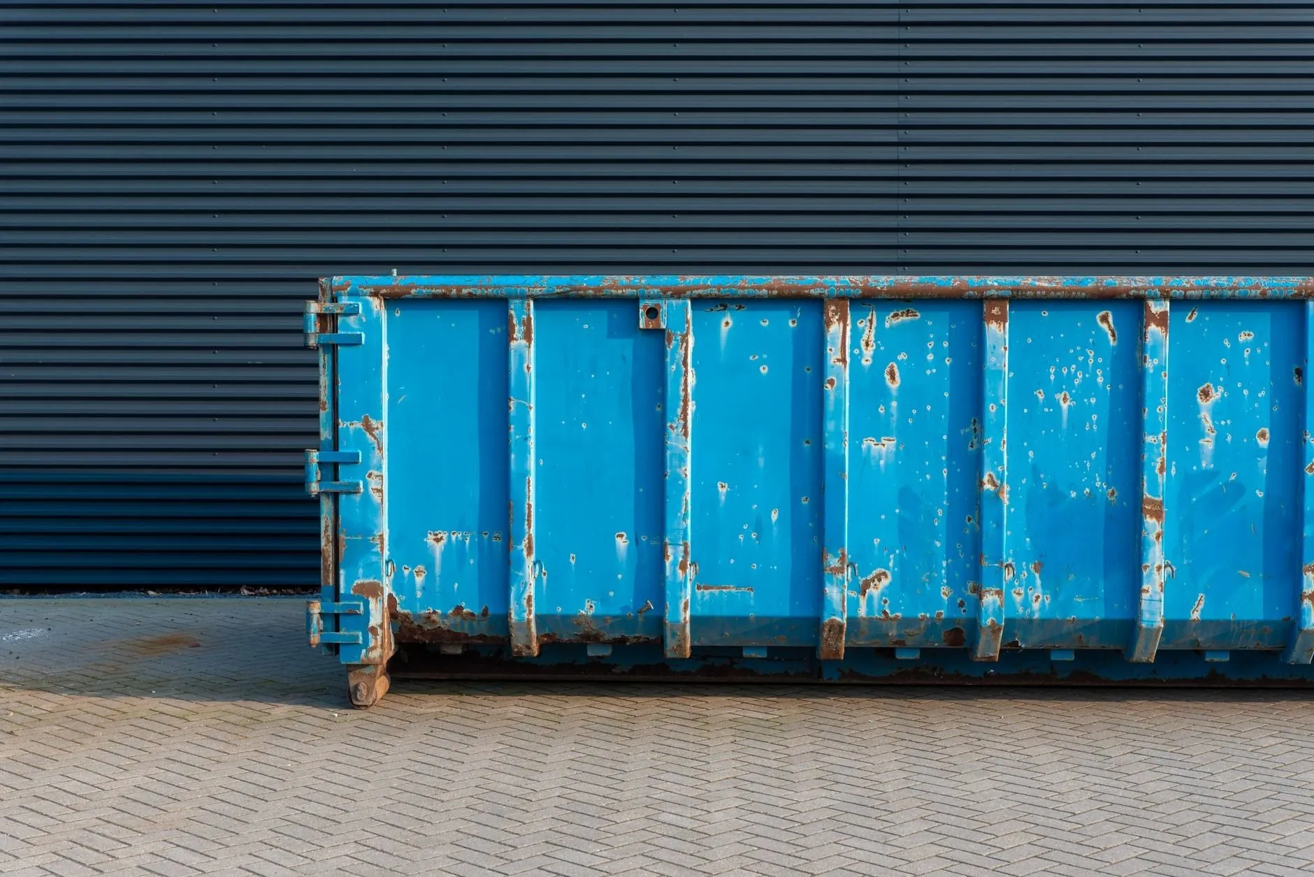 A weathered blue roll-on container with hook-lift rollers, standing against a dark ribbed wall