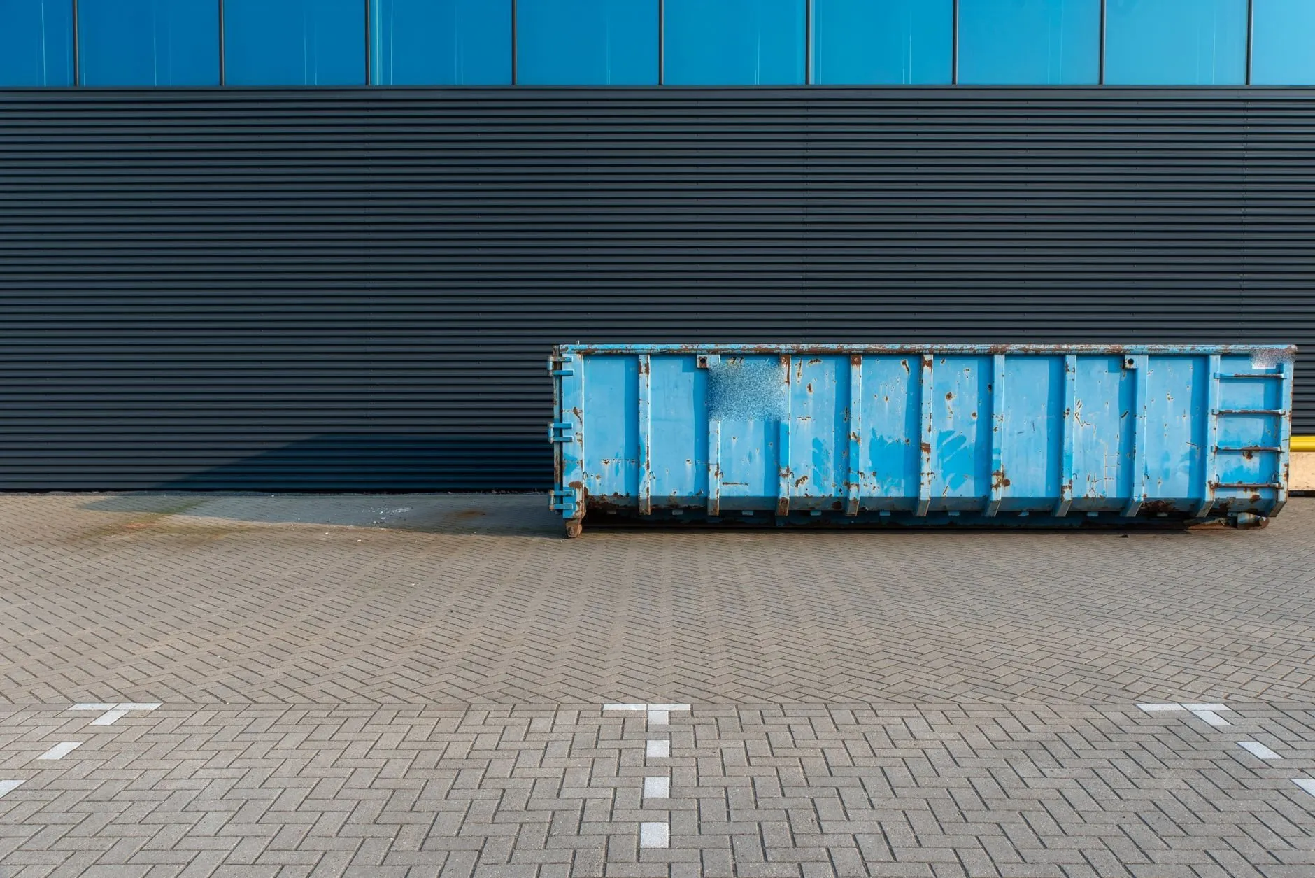 A long blue roll-on container standing on block paving in front of a dark clad warehouse