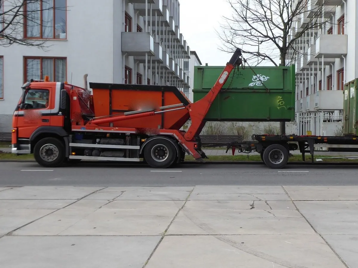 A skip lorry seen side-on on a street, its lifting arms raised, with a trailer of green containers behind