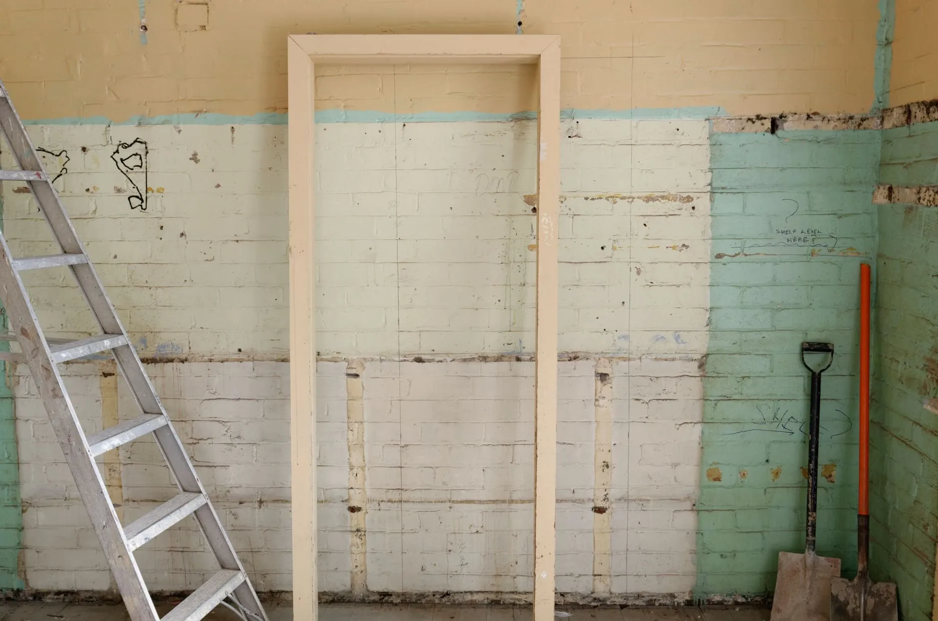 A stripped room with bare painted brickwork, an empty door frame, a stepladder and two spades